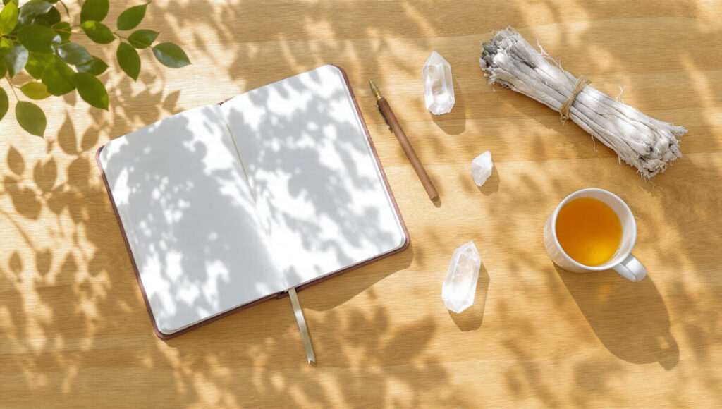 A sunlit flat lay featuring an open journal, clear quartz crystals, white sage, and tea on a wooden table, representing tools for spiritual healing rituals after a psychic reading for heartbreak.