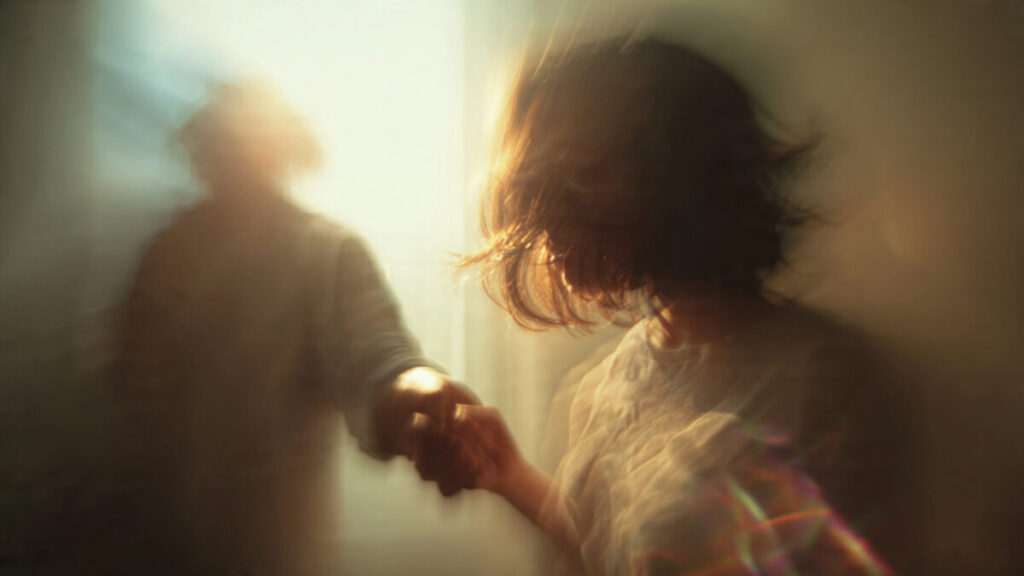 A dreamlike, blurry photograph showing a woman reaching towards an indistinct, faceless figure bathed in warm, golden light, symbolizing a spiritual connection in a dream.