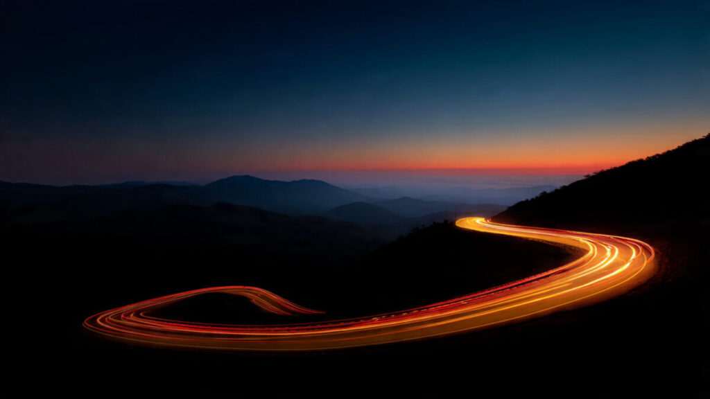 Long exposure photography of golden car light trails winding through a dark mountain landscape at twilight.