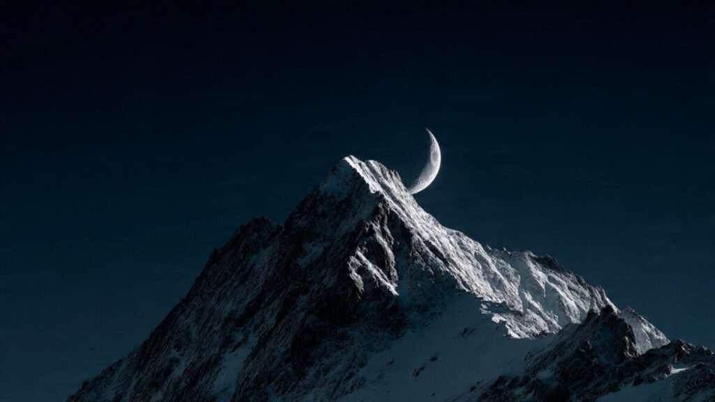 Low angle shot of a crescent moon rising over a sharp mountain peak.